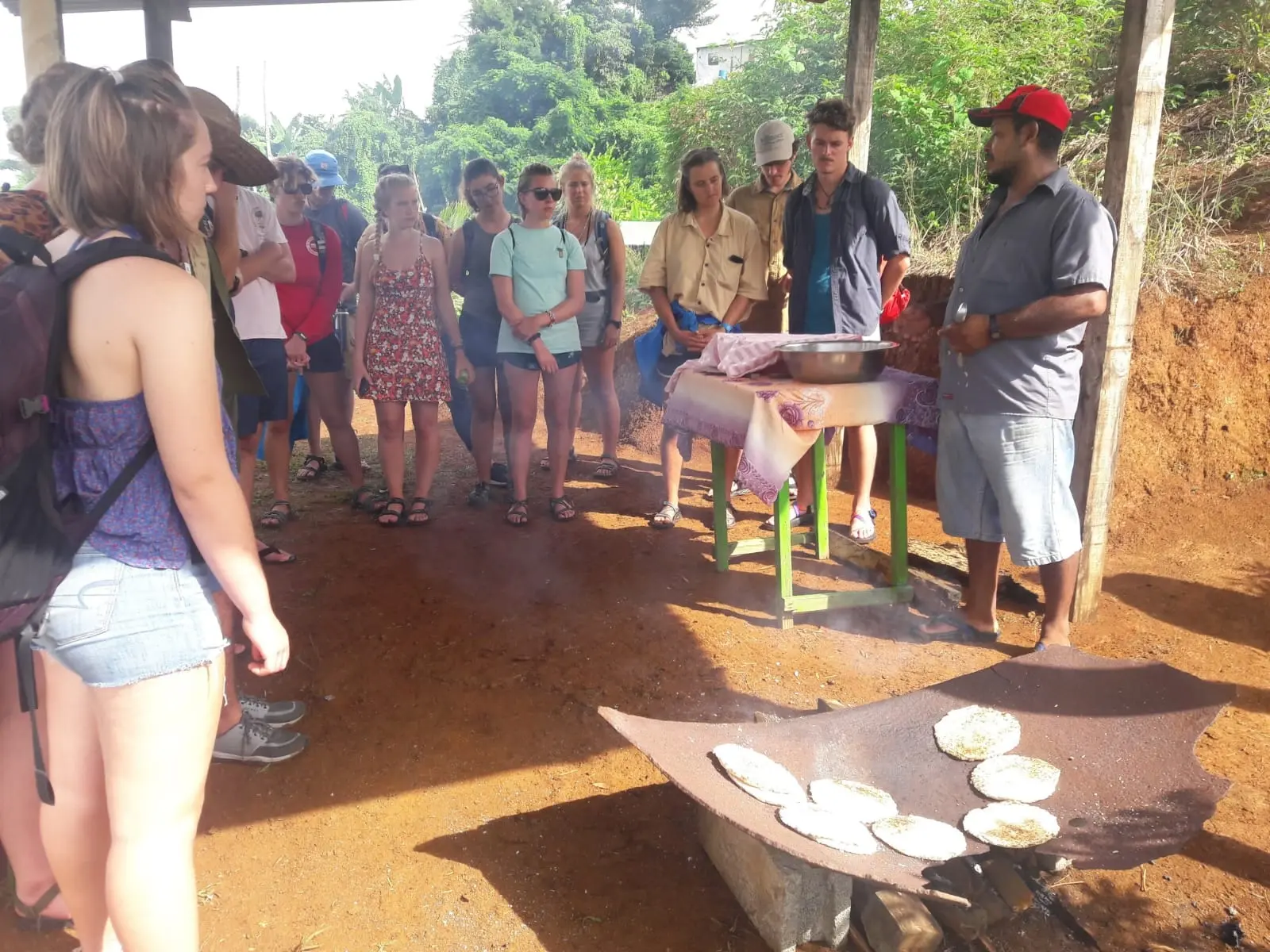 An outdoor scene showing a group of about a dozen young tourists standing in a semi-circle on a dirt floor under a wooden shelter. They are watching a man in a red baseball cap and grey shirt who is demonstrating how to cook several round flatbreads on a large, curved metal griddle heated by an open fire. Lush green tropical foliage and trees fill the background.