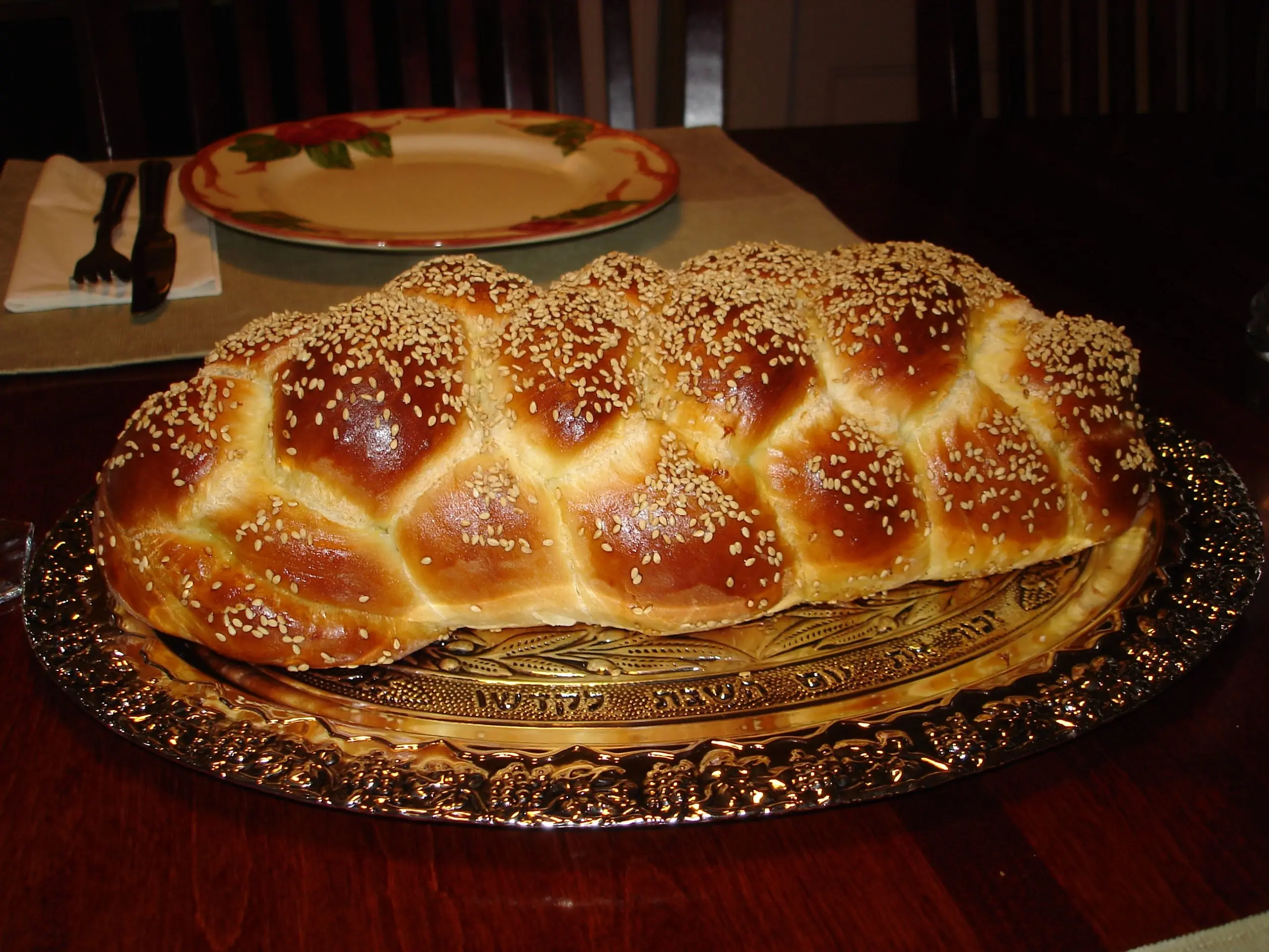 A large, beautifully braided challah bread with a shiny, golden-brown crust, generously topped with sesame seeds. The loaf is presented on an ornate silver-colored platter featuring etched designs and Hebrew text. In the blurred background, a dining table is set with a white and red decorative plate and silverware.