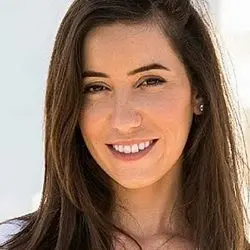 A close-up headshot of a smiling woman with long, dark brown hair, possibly the designer Danit Peleg, facing the camera outdoors in natural light.