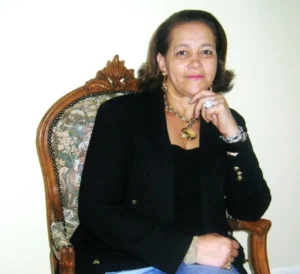 A middle-aged woman with brown hair, wearing a black blazer and a gold necklace, sitting in an ornate upholstered wooden chair. She is resting her chin on her hand and looking directly toward the camera.