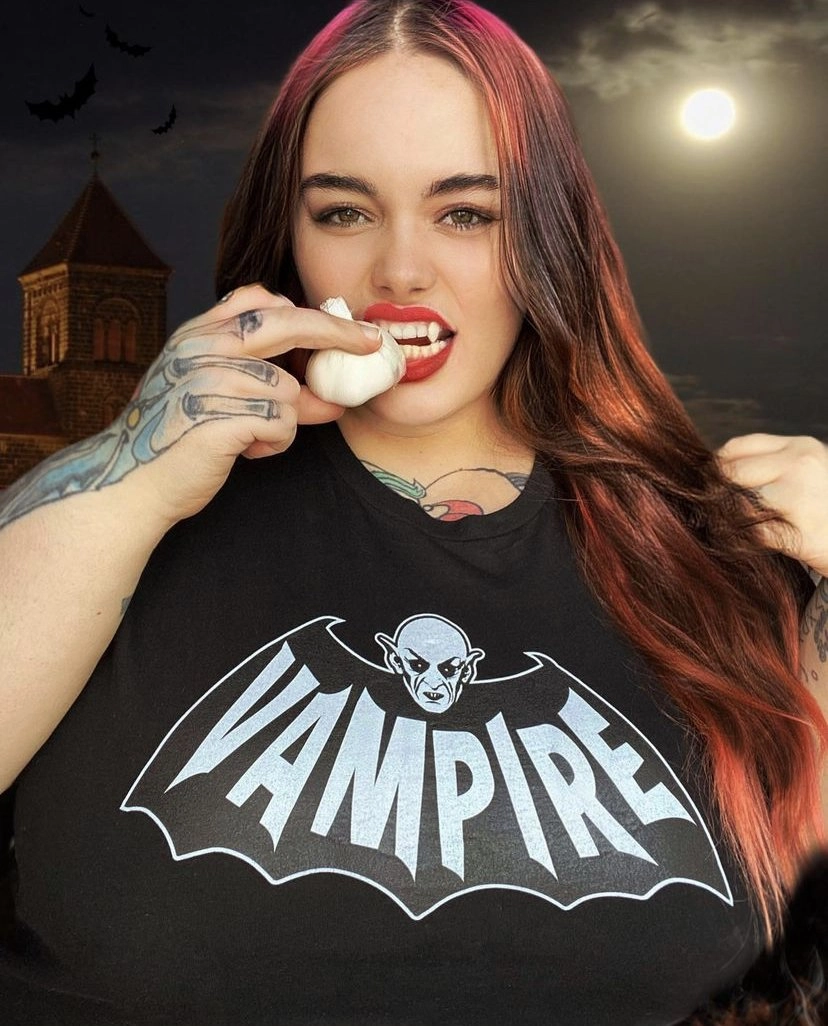 A close-up, digitally altered photo of a woman in a black t-shirt with a white 'VAMPIRE' logo featuring a bat and a vampire face. She is holding a bulb of garlic to her red-lipsticked mouth, which has visible vampire fangs. She has tattoos on her chest and arms. The background is a dramatic night scene with a full moon, a castle, and bats flying.