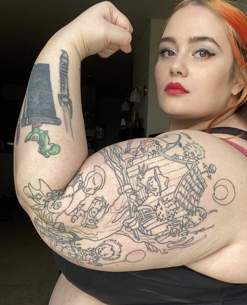 A portrait of a woman flexing her right bicep toward the camera, showing off a large, complex black-line tattoo on her upper arm that appears to depict cartoon characters on a boat in a stormy scene. She is wearing a black tank top and has red lipstick. Smaller tattoos, including a green creature and a sword, are visible higher up on her arm.