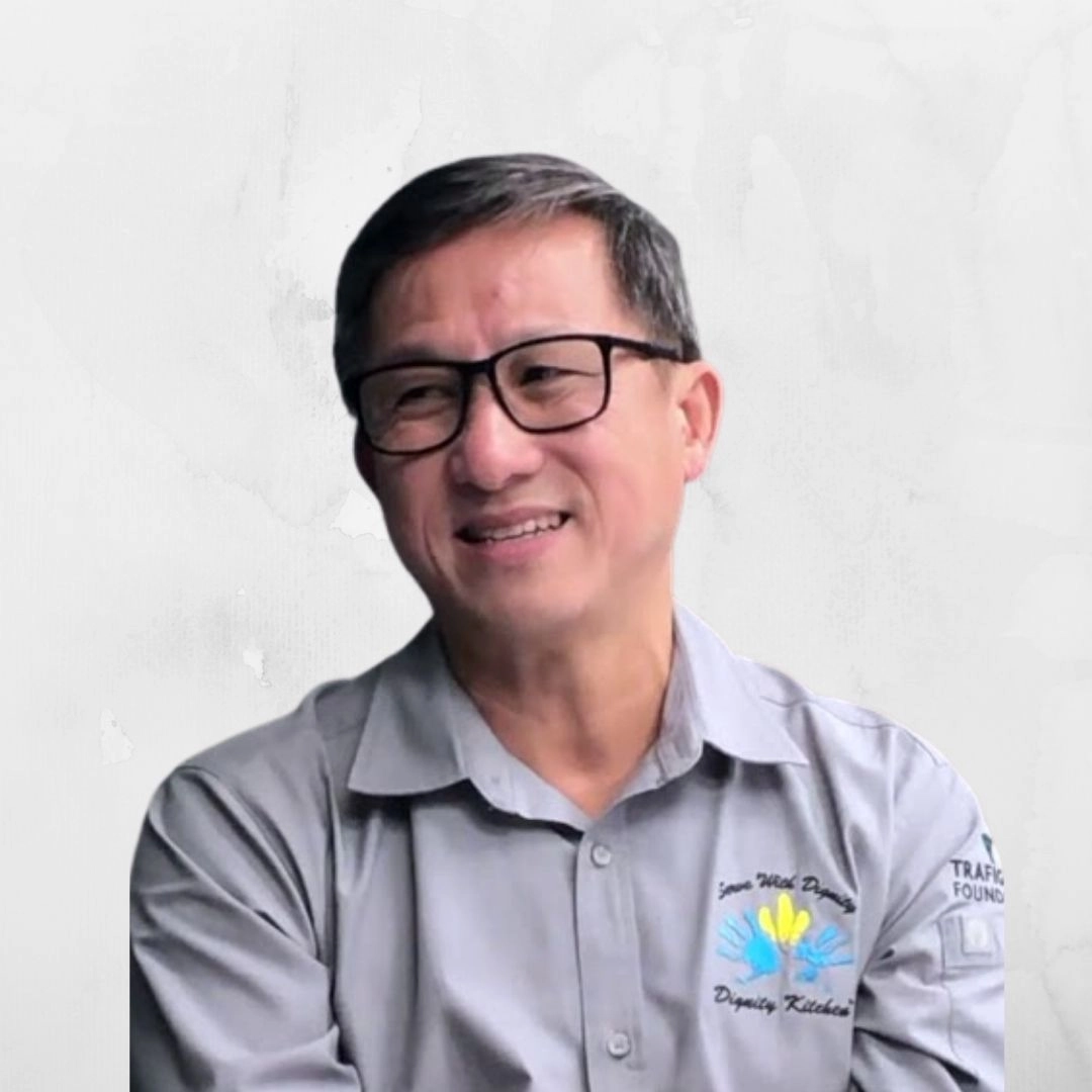 A medium shot of a middle-aged East Asian man with short dark hair and black-framed glasses, smiling and looking toward the left. He is wearing a grey button-down shirt that features a logo on the left chest with the words "Serve With Dignity" and "Dignity Kitchen" surrounding a graphic of blue and yellow handprints. The background is a plain, textured off-white wall.