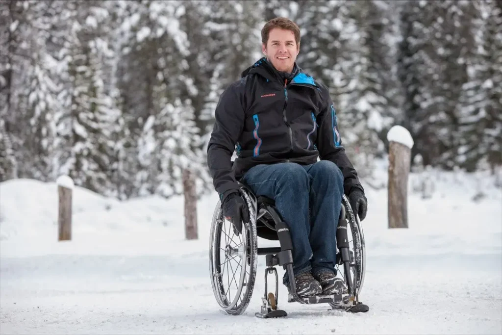 Among snow-covered pines a man sits in his wheelchair, wearing a winter jacket and a smile. The text reads, Patrick Mayer, Creator of Wheelblades “I decided to continue with my life and try to make the best of it”