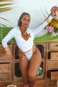 A close-up, waist-up shot of a woman in a long-sleeved, ruffled white one-piece swimsuit and gold jewelry, posing indoors against a decorated wooden cabinet.