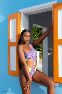 A woman posing in a one-shoulder, high-cut one-piece swimsuit featuring a colorful geometric print, a neon yellow/green panel, and a side cutout. She is leaning against a brightly painted blue wall with open orange shutters.