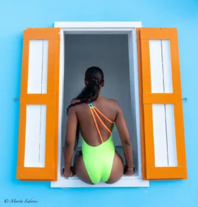 A rear view of a woman sitting on the sill of a small window, wearing a bright neon yellow/green, high-cut one-piece swimsuit with multiple neon orange and yellow straps across the open back. The window is set in a vibrant blue wall with bright orange shutters.