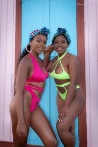 Two women pose side-by-side in bright swimwear. The woman on the left wears a hot pink strappy bikini, and the woman on the right wears a neon yellow/green strappy bikini. Both wear matching colorful headwraps, standing against a blue and pink paneled wooden background.