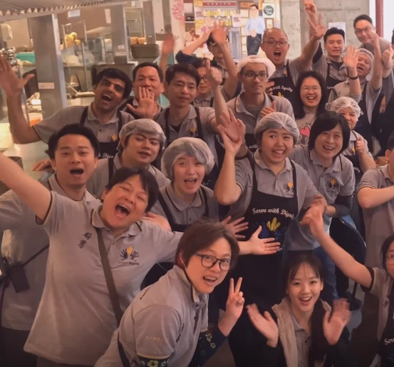 A joyful group portrait of a diverse team in a kitchen environment, showing about twenty people smiling and waving at the camera. Most are wearing uniform grey polo shirts, and several are wearing black aprons that say "Serve with Dignity" along with white hairnets. The group is densely packed, with several individuals making peace signs or cheering in a spirited atmosphere.
