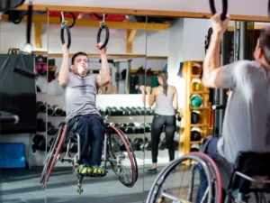A man in a wheelchair performs a pull-up using overhead gymnastics rings in a bright gym. He is wearing a grey t-shirt and dark pants, showing great focus and strength. His reflection is visible in a large wall mirror behind him, which also reveals other gym equipment like dumbbells and weight racks, as well as another person exercising in the background.