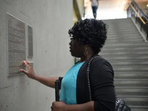 A woman with curly dark hair, wearing a teal top and black cardigan, stands in a hallway and uses her fingers to read a Braille sign mounted on a concrete wall. She holds a black blindness cane in her other hand. In the background, a wide concrete staircase leads upward, with another person visible in the distance at the top of the stairs.