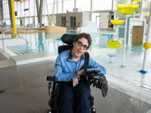 A young man with glasses and a blue jacket sits in a motorized wheelchair at the edge of a modern indoor swimming pool. The aquatic center features large floor-to-ceiling windows that let in ample natural light. In the background, there is a clear blue pool and a colorful water play area with yellow, flower-shaped fountains.