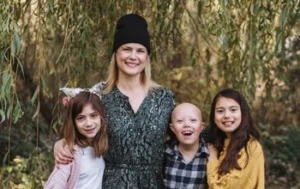 A group portrait of a smiling woman and three children standing outdoors in front of a large weeping willow tree. The woman, wearing a black beanie and a dark patterned dress, has her arms around two of the children. To her left is a young girl in a pink cardigan and white skirt; to her right is a young boy with alopecia wearing a plaid shirt, and next to him is an older girl in a yellow sweater and jeans. The ground is covered in fallen leaves and green grass.