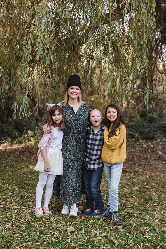 A group portrait of a smiling woman and three children standing outdoors in front of a large weeping willow tree. The woman, wearing a black beanie and a dark patterned dress, has her arms around two of the children. To her left is a young girl in a pink cardigan and white skirt; to her right is a young boy with alopecia wearing a plaid shirt, and next to him is an older girl in a yellow sweater and jeans. The ground is covered in fallen leaves and green grass.