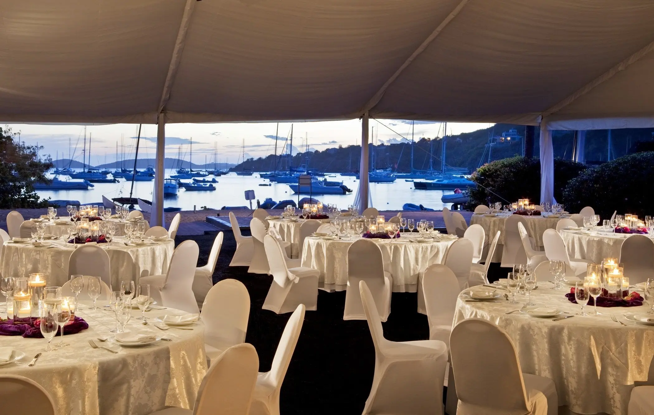 A formal outdoor dinner setting under a large white tent with a view of a harbor filled with anchored boats and yachts at twilight. The tables are round, covered in white linens, and set with white chairs, glassware, plates, and lit candles.