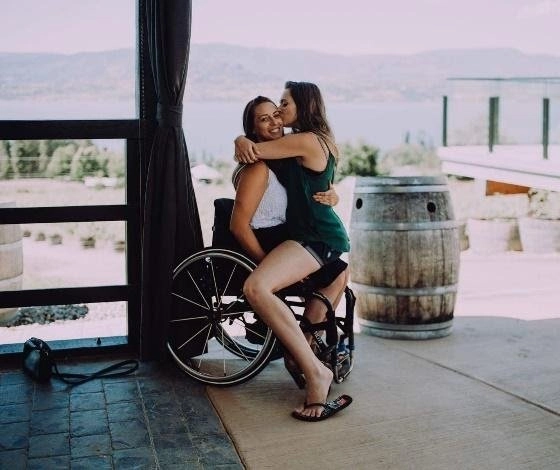 Two young women embrace on a covered patio with a large lake and mountains visible in the background. The woman on the left is seated in a wheelchair and is being hugged from behind by the second woman, who is sitting on her lap and kissing her cheek. A large wooden wine barrel is visible to the right.