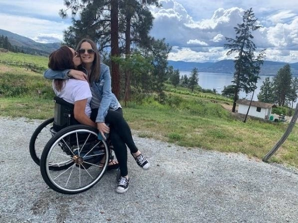 Two young women embrace while sitting on a gravel path overlooking a large lake and vineyards on a cloudy day. The woman on the left is seated in a wheelchair, wearing a white shirt and a beanie, while the woman sitting on her lap is wearing a denim jacket and black sneakers. The woman on the lap is kissing the other woman on the cheek.