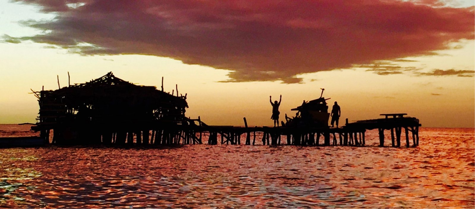 In a cloudy sky, the sun sets over copper-hued water. In silhouette, two people stand on a ramshackle dock, one with her arms raised to the sky.