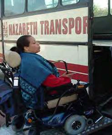A side view of a person seated in a dark blue motorized mobility scooter, preparing to board a bus. The individual is wearing a red top and a blue patterned shawl or blanket over their shoulders. They are positioned on a lift or ramp that appears to be integrated into the doorway of a large white bus. The side of the bus above them is visible and features the word "NAZARETH TRANSPOR" in large, red lettering.