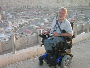A middle-aged man with a short white beard wearing a light-colored shirt and blue jeans is seated in a black and blue motorized wheelchair. He is holding a camera on his lap and looking directly at the viewer with a neutral expression. He is positioned on a raised, paved area with a low stone railing. In the background, there is a sprawling, detailed architectural model of an ancient city, possibly Jerusalem, with many small, densely packed stone-colored buildings under bright daylight.