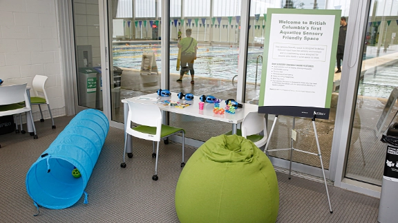 A playroom, with a giant blue tube and large green beanbag, chairs and table holding bright objects has a glass wall through which we see a large swimming pool. The article is titled Accessible Metro Vancouver.