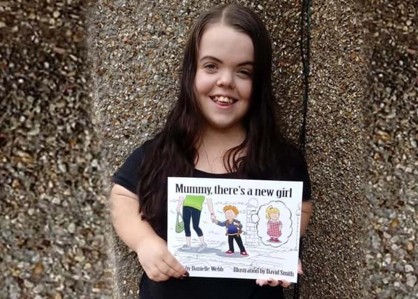 A smiling woman stands against a stucco wall, holding a brightly colored publication entitled, Mommy there's a new girl. . .The headline reads, Danielle Webb on Dwarfism.