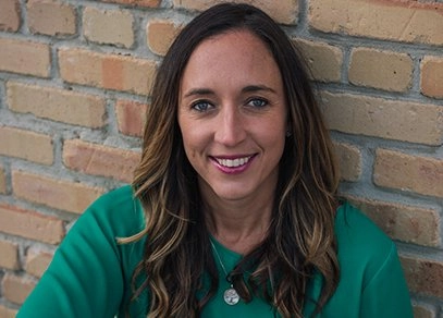 A close-up photograph of a woman with long, brown, wavy hair and blue eyes, smiling directly at the camera. She is wearing a bright green top and a silver necklace with a circular pendant, and is leaning against a rustic, light brown brick wall.