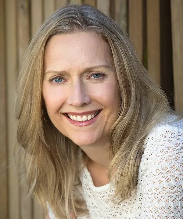 A headshot of a smiling Blonde woman with blue eyes, wearing a white crocheted or lace top. Her long hair is parted in the middle and falls over her shoulders. She is posed outdoors in front of a vertical wooden slat fence or wall.