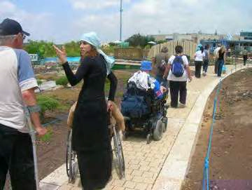 A group of people walking outdoors on a paved path surrounded by dry earth and some low vegetation. In the foreground, a woman wearing a long black dress and a light blue head covering is pushing herself in a manual wheelchair. She is pointing with her right hand towards the background, where a man is walking with crutches. Further along the path, a person is seated in a motorized wheelchair followed by several other people, some wearing backpacks, walking away from the camera. The background has some distant modern buildings and structures.