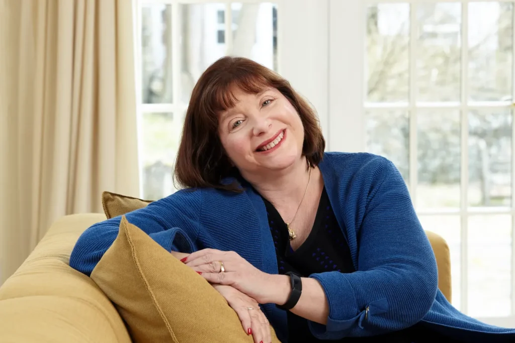 A smiling woman in a blue dress sits on a sofa in a bright room. White French doors and beige drapes are behind her. Traveling Different is the headline, followed by: 5 questions for author, Dawn Barclay, Traveling Different. Vacation Strategies. . . Read More.