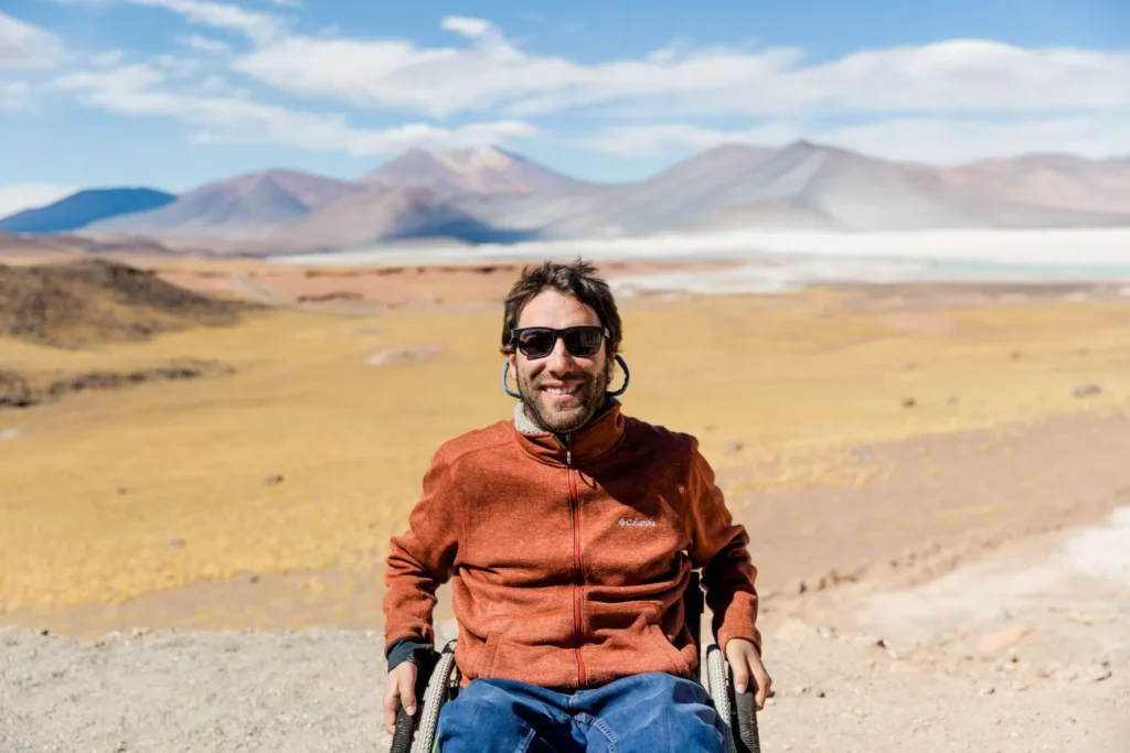 A man wearing casual clothes, sunglasses and a smile sits in a wheelchair. Behind him is an expanse of arid land, with mountains rising in the distance under a blue sky studded with fluffy clouds. Overlaying text reads, Wheel the World, We made the impossible possible. If there's one thing you should know about Camilo Navarro and Avaro Silberstein. . . an orange button says Read More.