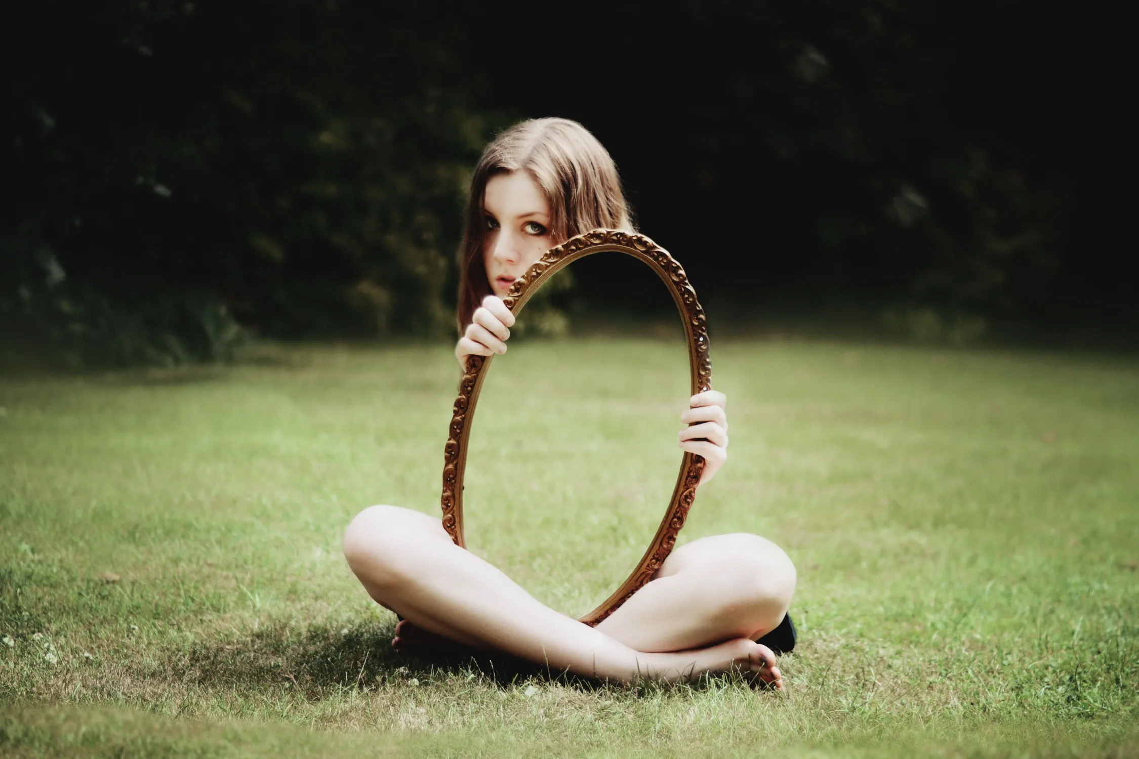 A girl sits cross-legged on the grass, holding a large oval mirror that reflects the grass, making her body appear invisible. Text reads, When Invisible Disabilities Reveal Themselves, by Lisa Guthrie Deabill. Read More.