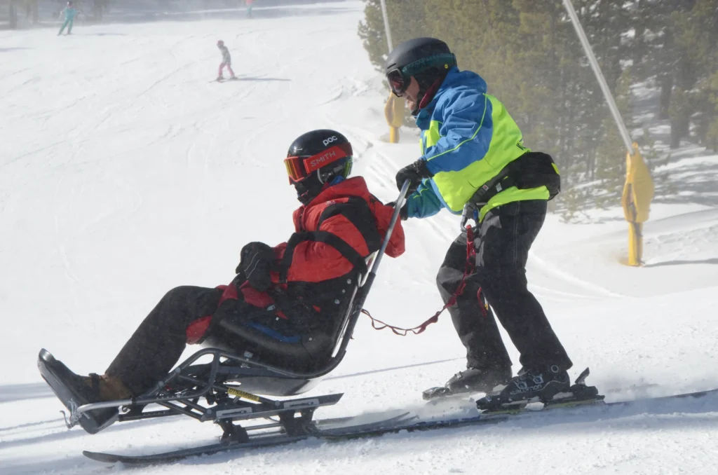 On a snowy ski hill a skiier in a neon green jacket pushes an adapted wheelchair skiier in a red jacket. The headline reads, Accessible travel is hard. . . By Josh Basile, C4-5 Quadriplegic, Trial Attorney, Disability Rights Advocate. Orange text says, Read More.
