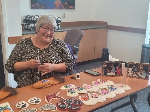 Smiling woman seated at a table displaying various handcrafted items, including mittens and fabric patches decorated with floral and paw print designs. The table also has framed photos and sewing materials. She appears to be working on one of the pieces, and the setting is an indoor room with neutral decor and a few chairs in the background.
