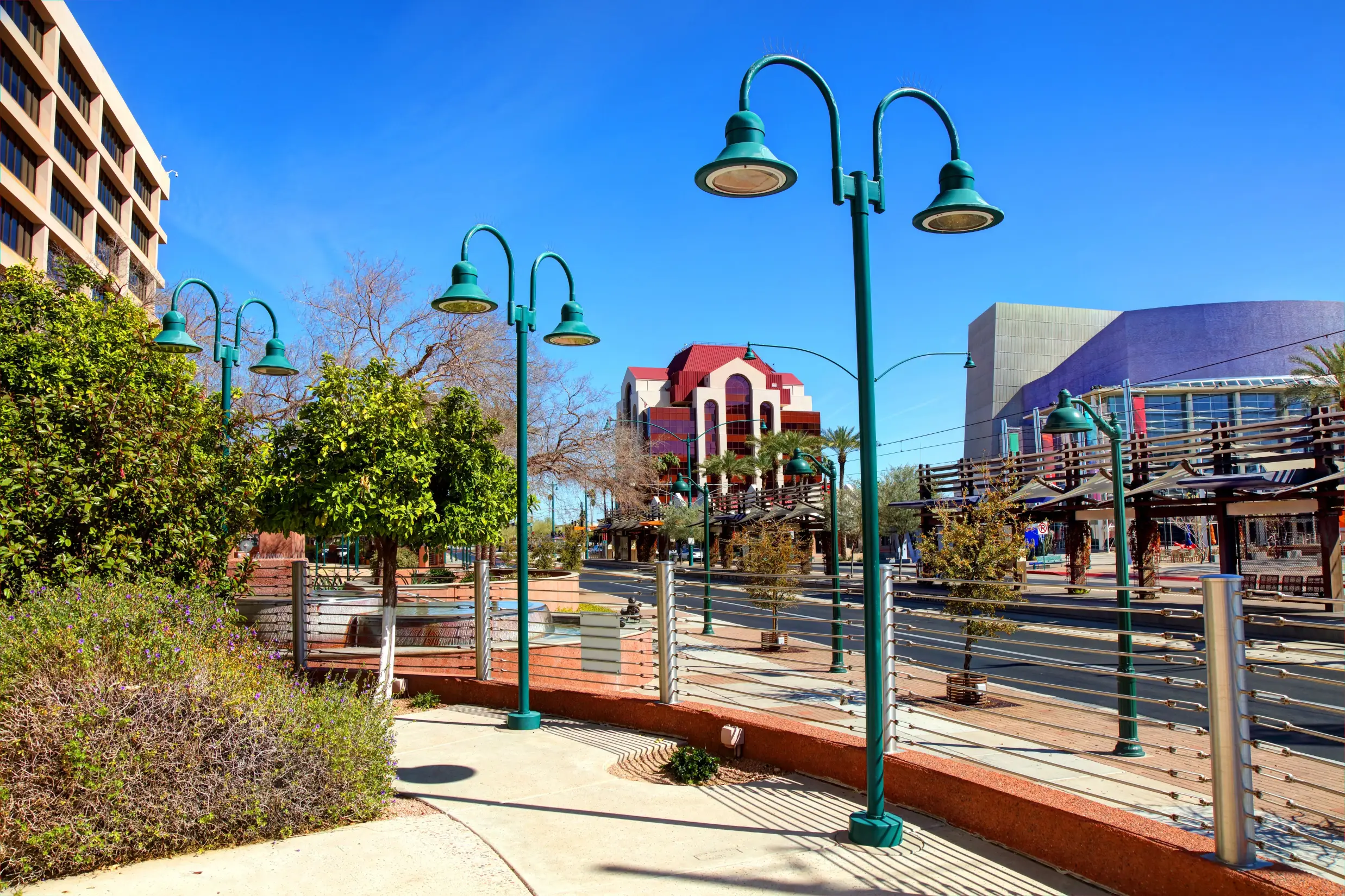 A sunny daytime view of a pedestrian walkway in an urban area, possibly near Scottsdale, Arizona, lined with distinctive tall lampposts featuring curved arms and teal-colored lights. In the background, a street runs parallel to the path, and buildings of varied architecture are visible, including a prominent structure with a terracotta roof and a modern building with a large curved blue facade. Green trees and low shrubs frame the walkway.