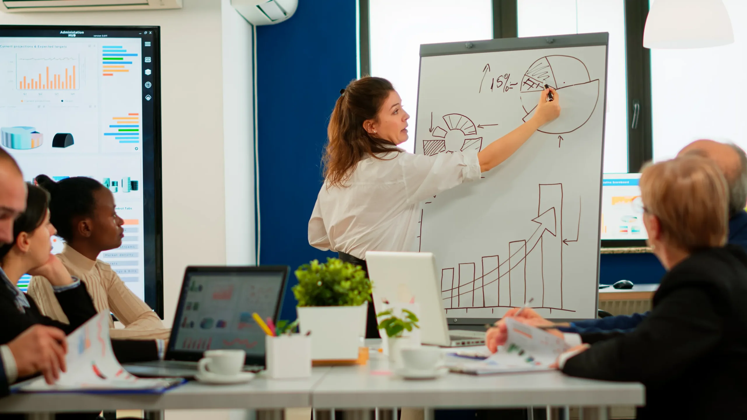 In a bright room, a woman holds a marker to a whiteboard full of graphs and pie charts. Her mouth is open, mid-speech as a group of people watch her from the nearby table, their laptops open in front of them.
