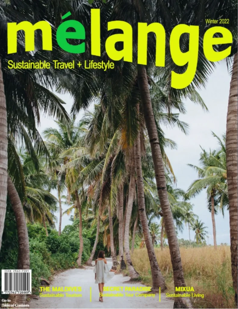 The cover features a vibrant tropical scene with a lush pathway lined by tall, green palm trees. A woman in a white dress walks down the path, adding a serene and exploratory vibe. The title "mélange" is prominently displayed at the top in bold, bright yellow and green letters, with the subtitle "Sustainable Travel + Lifestyle" just below it. This edition highlights sustainable tourism in the Maldives, a secret paradise, and sustainable living tips. The overall aesthetic promotes a theme of eco-friendly travel and lifestyle choices.