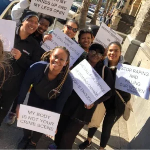 A group of smiling women of diverse backgrounds standing close together in an outdoor urban setting, holding up protest signs advocating for an end to gender-based violence and femicide. Signs include text such as "WE CALL FOR JUSTICE," "STOP RAPING AND KILLING OUR WOMEN," and "MY BODY IS NOT YOUR CRIME SCENE."