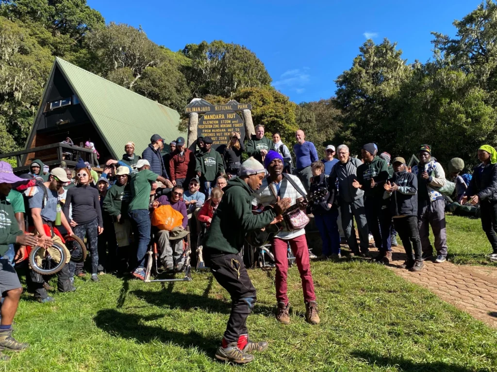 A large group of hikers, including people in wheelchairs and adaptive bikes, gathered in a grassy area outside a wooden cabin with a green roof, marked as the "Mandara Hut" in Kilimanjaro National Park. Two people in the foreground are interacting, and the group is standing in front of a wooden sign in a mountainous, forested environment under a bright, sunny sky.