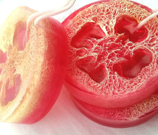 A close-up, high-detail photo of several round bars of soap, which appear to be pink or reddish-orange, each containing a slice of natural loofah sponge embedded inside. The loofah's fibrous texture and the clear, vibrant soap form a pattern. The bars have a white string or loop attached, suggesting they are meant for hanging in the shower.