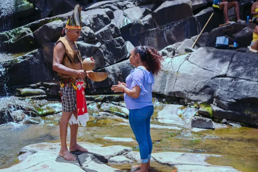 An indigenous man and a woman stand barefoot facing each other on wet rocks near a small waterfall, possibly participating in a ceremony. The man, on the left, wears a traditional feathered headdress, a fringed leather vest, and a plaid skirt with a white and red sash, and he holds two carved coconut or wooden bowls. The woman, on the right, wears a lavender top and blue jeans, with her hands together. Large, dark, jagged rocks form the background.