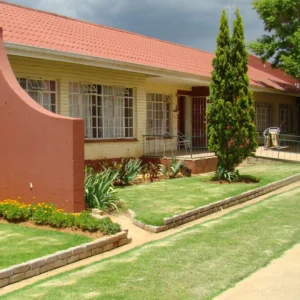 The exterior of a single-story, light yellow building with a red tile roof, likely a residential home or small community center, set against a cloudy sky. The front yard is well-maintained with a green lawn, garden beds, and a tall, narrow cypress tree.