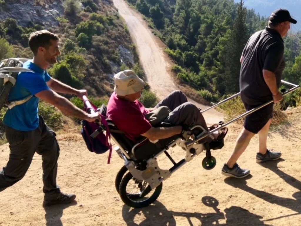 Two men are hiking up a steep, dirt path surrounded by green forest, pushing and pulling a third person seated in a specialized, single-wheeled off-road wheelchair. The person in the chair is wearing a red shirt and a wide-brimmed sun hat. The man in front wears a blue shirt and a backpack, and the man in the back wears a dark gray shirt.