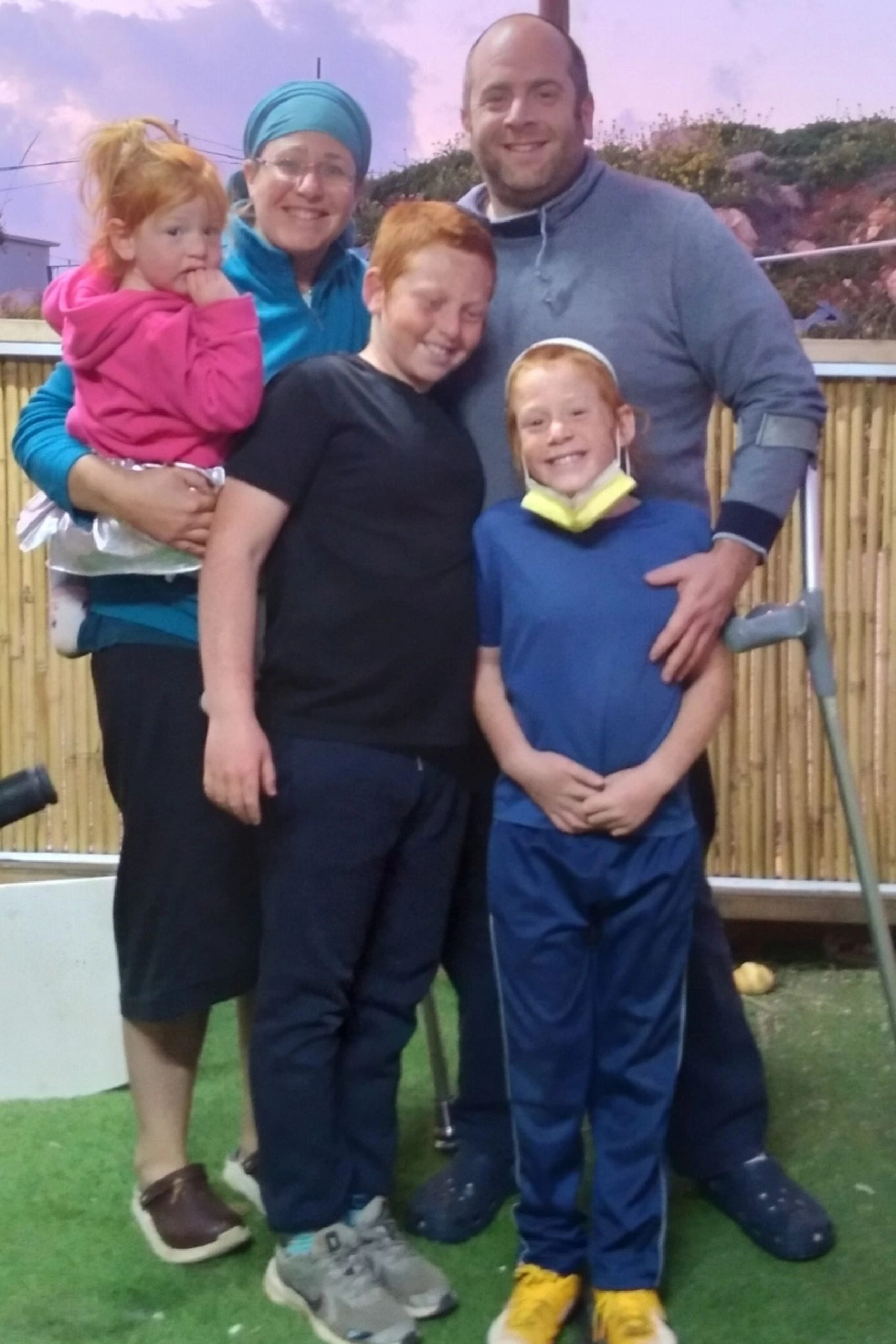 A family portrait showing two adults and three children posing outdoors in the evening. The mother, wearing a blue head covering and jacket, holds a small girl in a pink top. The father, wearing a gray sweatshirt, stands with his arm around a boy and a girl, both with bright red hair and wide smiles. A crutch or walking aid is visible by the father's right leg.