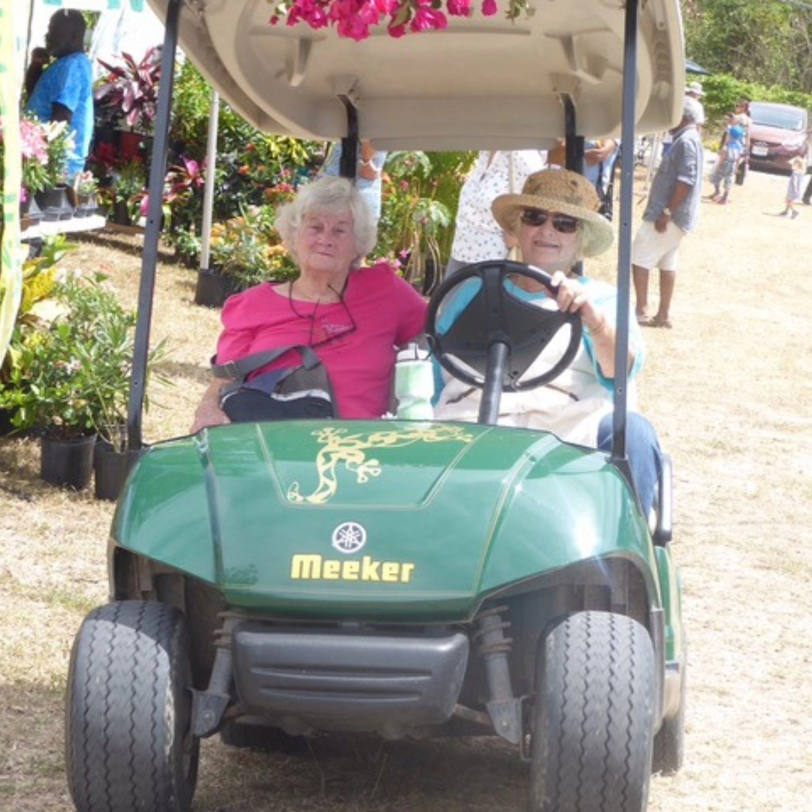 Two elderly women are seated in a green utility golf cart at an outdoor event, possibly a fair or market. The woman in the driver's seat wears a straw sun hat and sunglasses, and the woman next to her wears a bright pink shirt. The cart has the name "Meeker" on the front.