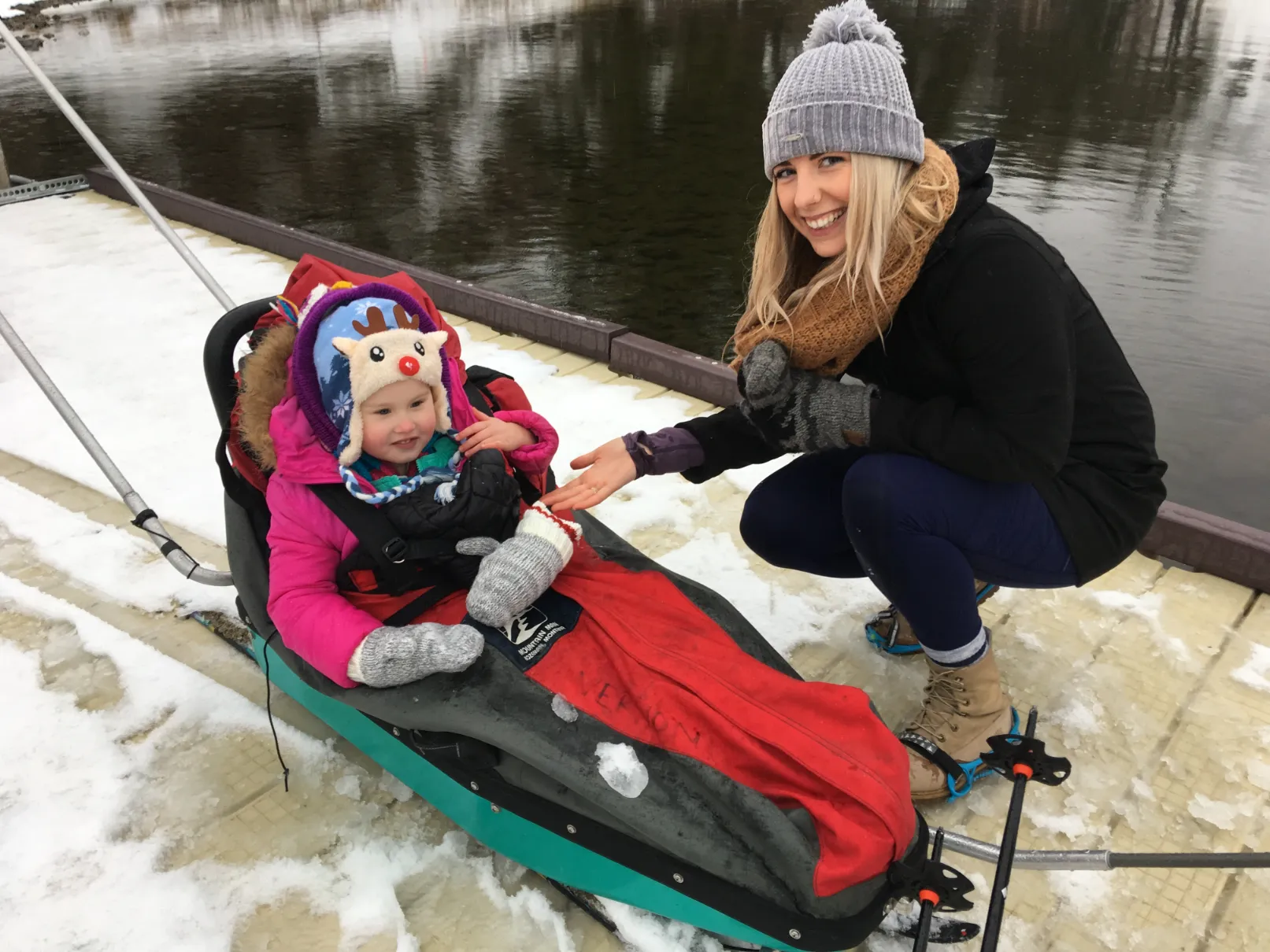 A young woman with blonde hair, wearing a grey knit hat and a brown scarf, is kneeling down next to a small child who is seated in an adaptive sled. The child is bundled in a pink snowsuit, mittens, and a whimsical winter hat, smiling at the woman who is gently reaching out a gloved hand. They are situated on a snowy, wooden dock next to dark water.
