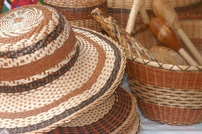 A close-up shot of a variety of woven indigenous handicrafts. In the foreground are two wide-brimmed hats with a repeating pattern of light tan, brown, and black weaving. To the right, a large woven basket contains wooden tools and musical instruments, including a maraca.
