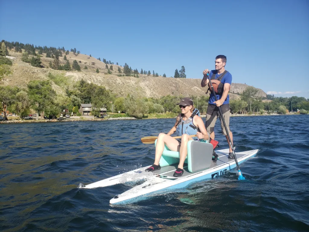 An adapted paddleboard on a lake holds two people; one sits in a built-in seat and paddles, one stands behind and paddles. They pass by dry rocky hills.