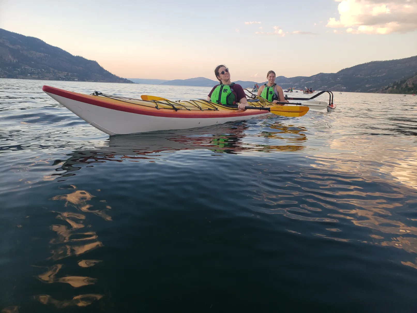 Two people wearing bright green life vests are paddling a red and yellow tandem sea kayak on a large, calm body of water during the evening. Both are smiling and looking toward the camera. One person wears glasses and has short dark hair. Steep, forested hills rise directly from the water in the background under a pale orange and blue sky.