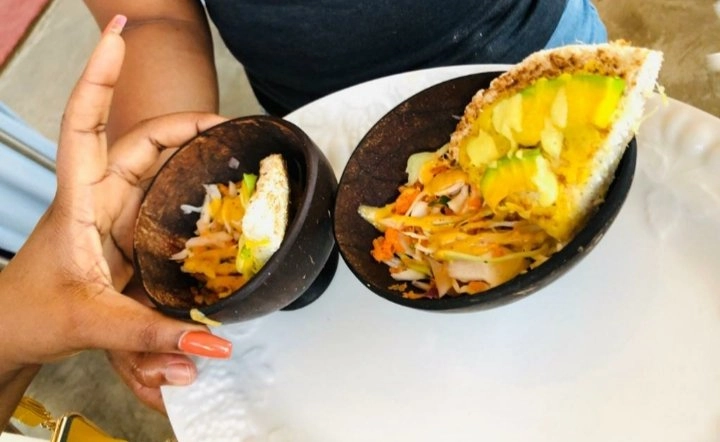 A person's hand with orange-tipped nails holds a white plate containing two small, dark brown bowls made from coconut shells. Each bowl holds a salad or slaw topped with a triangular piece of bread or flatbread, garnished with sliced avocado and drizzled with an orange-yellow sauce.